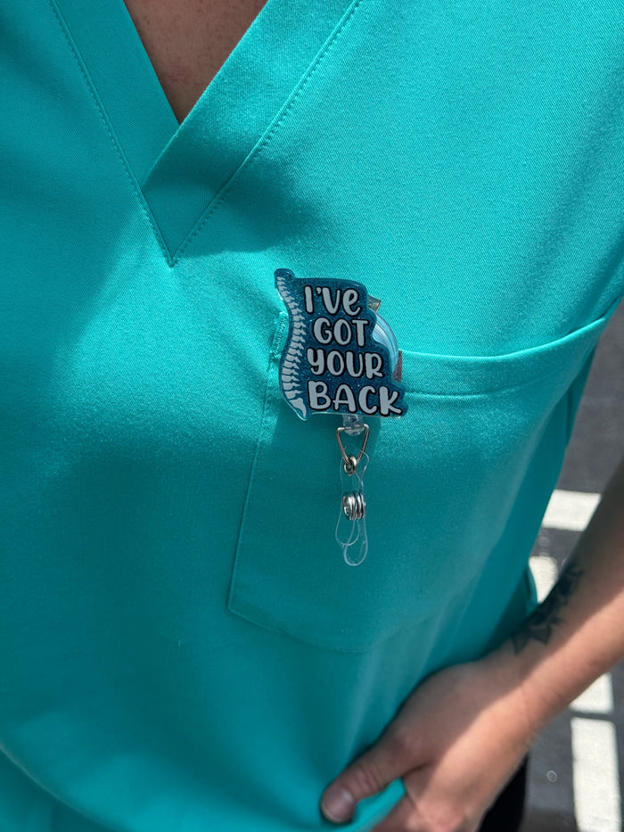 Person with Blue Glitter I'VE GOT YOUR BACK Spine Badge Reel clipped on front pocket of scrub shirt