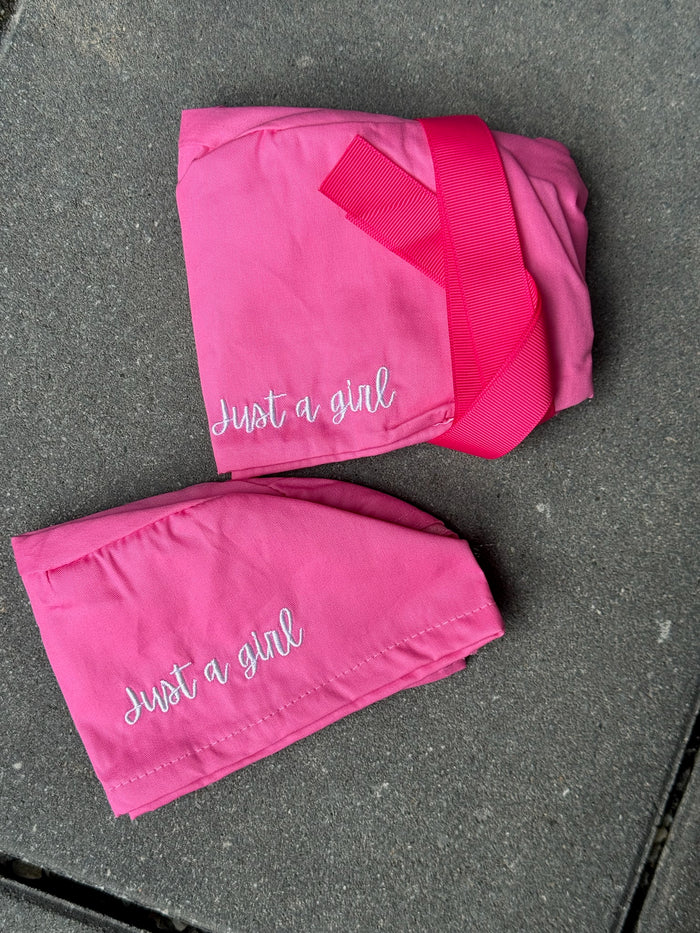 Two pink pairs of scrub hats with 'Just a girl' embroidered text on a gray surface