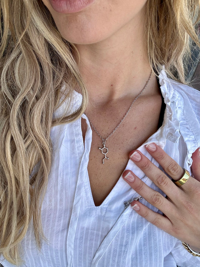Close-up of a person wearing a white blouse with a silver Noradrenaline Molecule necklace and gold ring.