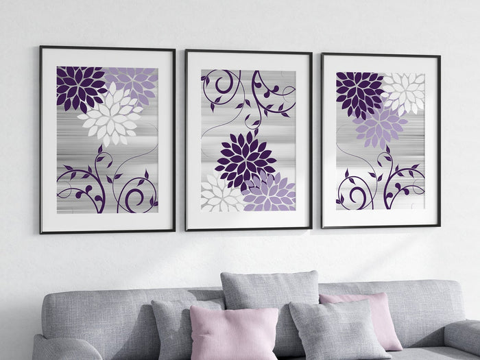 A set of three purple and gray floral wall art prints arranged vertically on a wall above a gray sofa with pink and gray cushions.