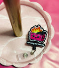 Retractable badge holder with a cartoon dumpster on fire design and 'surviving not thriving' text, in a pink tray